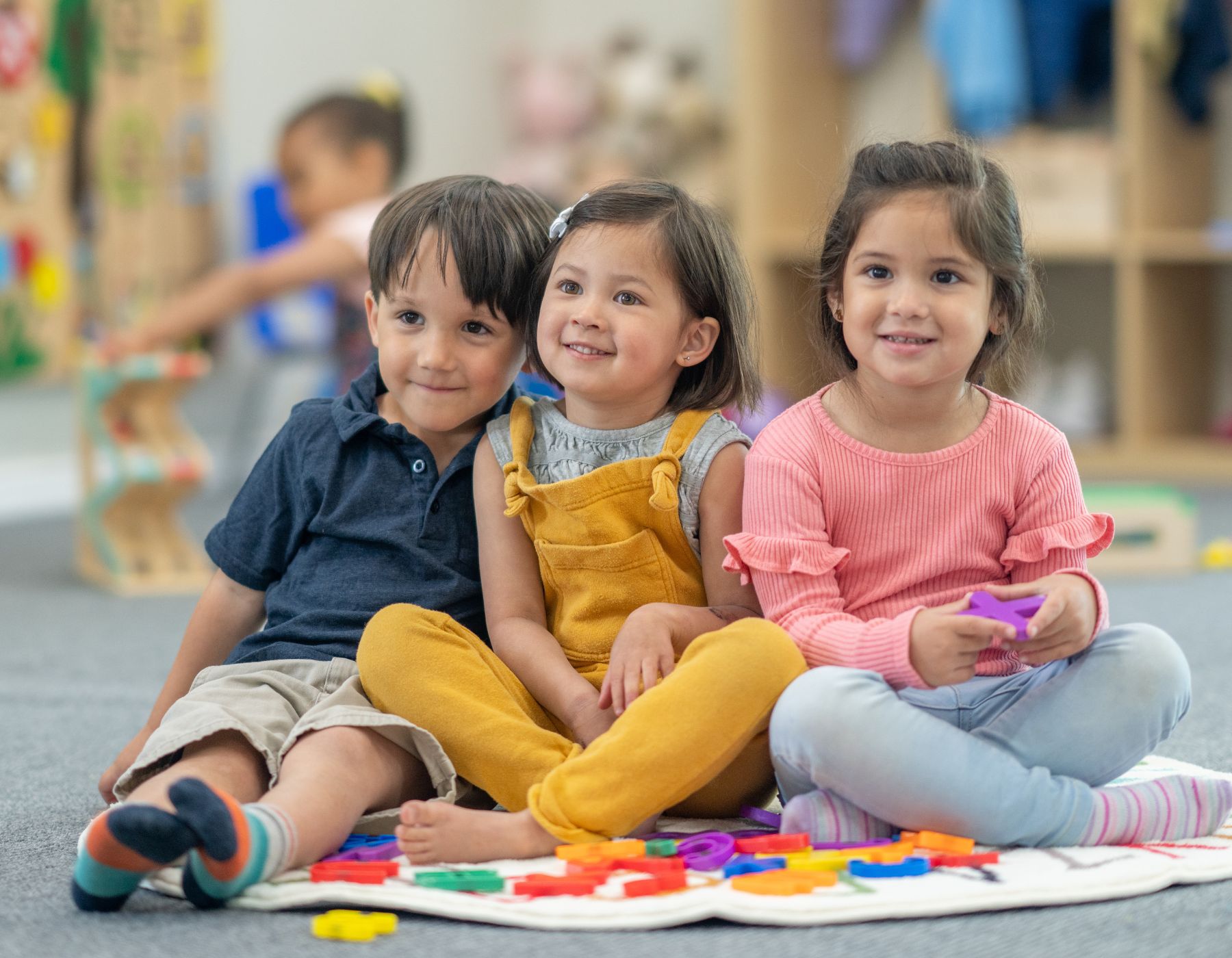 Tres niños pequeños de diferentes etnias sentados en el suelo de un aula de kinder, sonriendo y jugando con bloques de construcción de colores.
